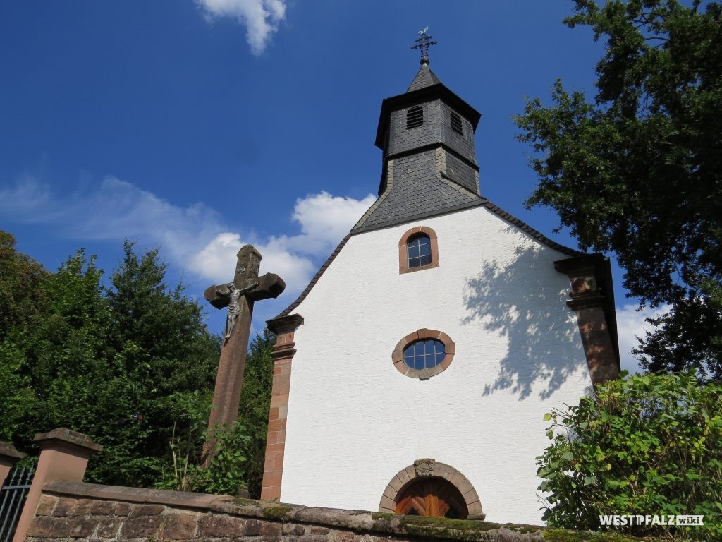 Westansicht der Rochuskapelle in Kaiserslautern Hohenecken. Links daneben ist eine Jesusfigur an einem großen Steinkreuz zu erkennen.