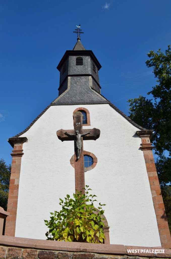 Westansicht der Rochuskapelle in Kaiserslautern Hohenecken. Links daneben ist eine Jesusfigur an einem großen Steinkreuz zu erkennen.