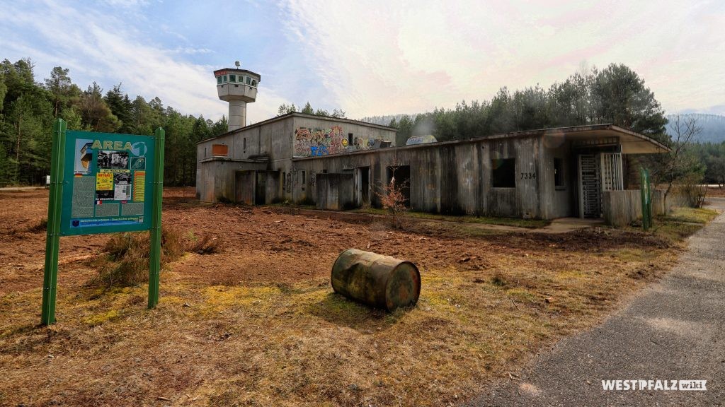 Ehemaliges US-Sonderwaffenlager Area One in Fischbach bei Dahn. Blick auf eines der heute leerstehenden Gebäude, angrenzend an den Tower. Im Vordergrund eine moderne Informationstafel sowie ein altes Metallfass.