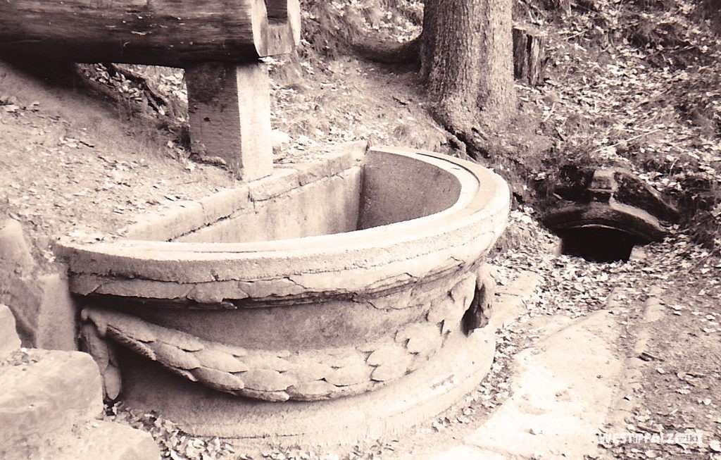 Die Hälfte der Brunnenschale aus dem Garten des Trippstadter Schlosses als Fassung für den Wasserablauf des Burgalbweihers. Das Wasser fließt über eine Zuleitung aus Holz in das Auffangbecken.