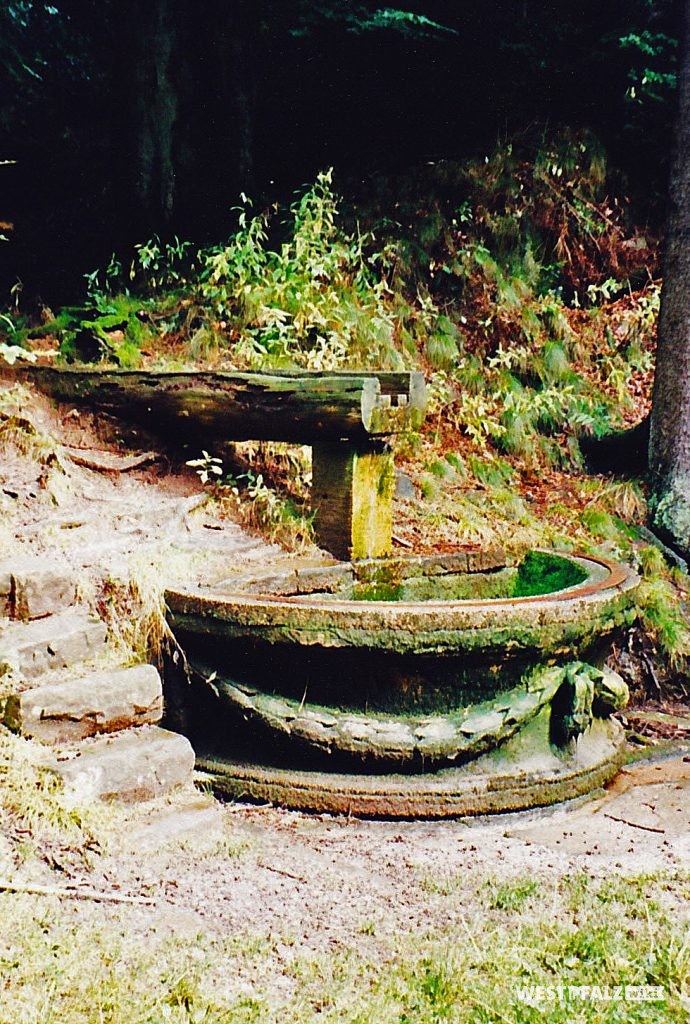 Die Hälfte der Brunnenschale aus dem Garten des Trippstadter Schlosses als Fassung für den Wasserablauf des Burgalbweihers. Das Wasser fließt über eine Zuleitung aus Holz in das Auffangbecken.